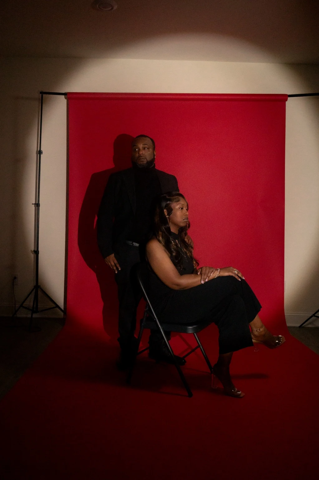 A behind-the-scenes studio portrait of a couple in black clothing on a red seamless backdrop, with the studio setup visible.