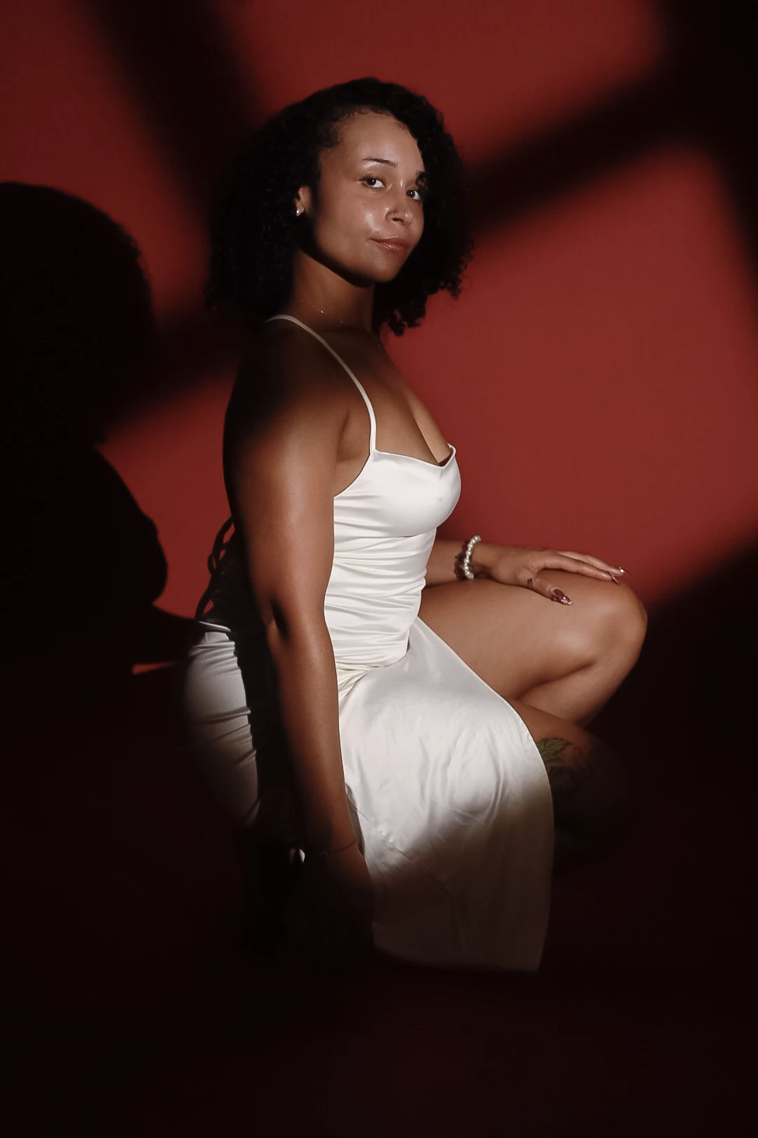 Editorial portrait of a woman in a satin dress against a crimson backdrop with dramatic window-shaped shadows.